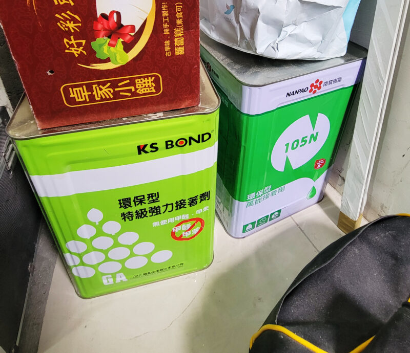 施工現場並排的兩罐接著劑，罐標為KS BOND環保型特級強力接著劑，標示無使用甲醛、甲苯，以及南寶105N環保型萬能接著劑