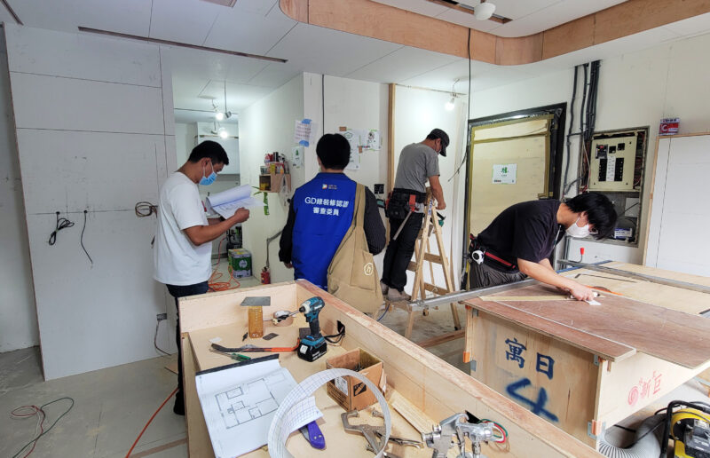 施工中的室內現場，身穿GD綠裝修認證審查委員背心的人員與手持圖面的現場人員、木作工班同在工地