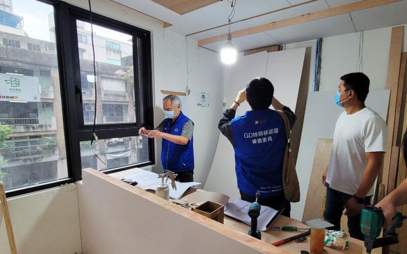 施工中的窗邊貼有保持通風標示，牆面貼有建材堆放整齊標示，審查委員背心人員與工班在現場查看