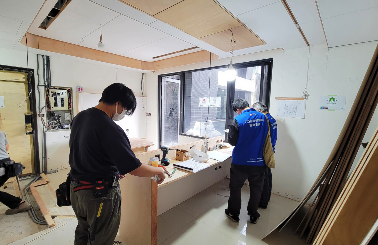室內設計公司施工中的工程管理現場，審查委員在窗邊對照圖面查看，木作師傅在一旁丈量