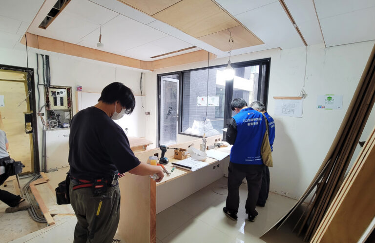 室內設計公司施工中的工程管理現場，審查委員在窗邊對照圖面查看，木作師傅在一旁丈量